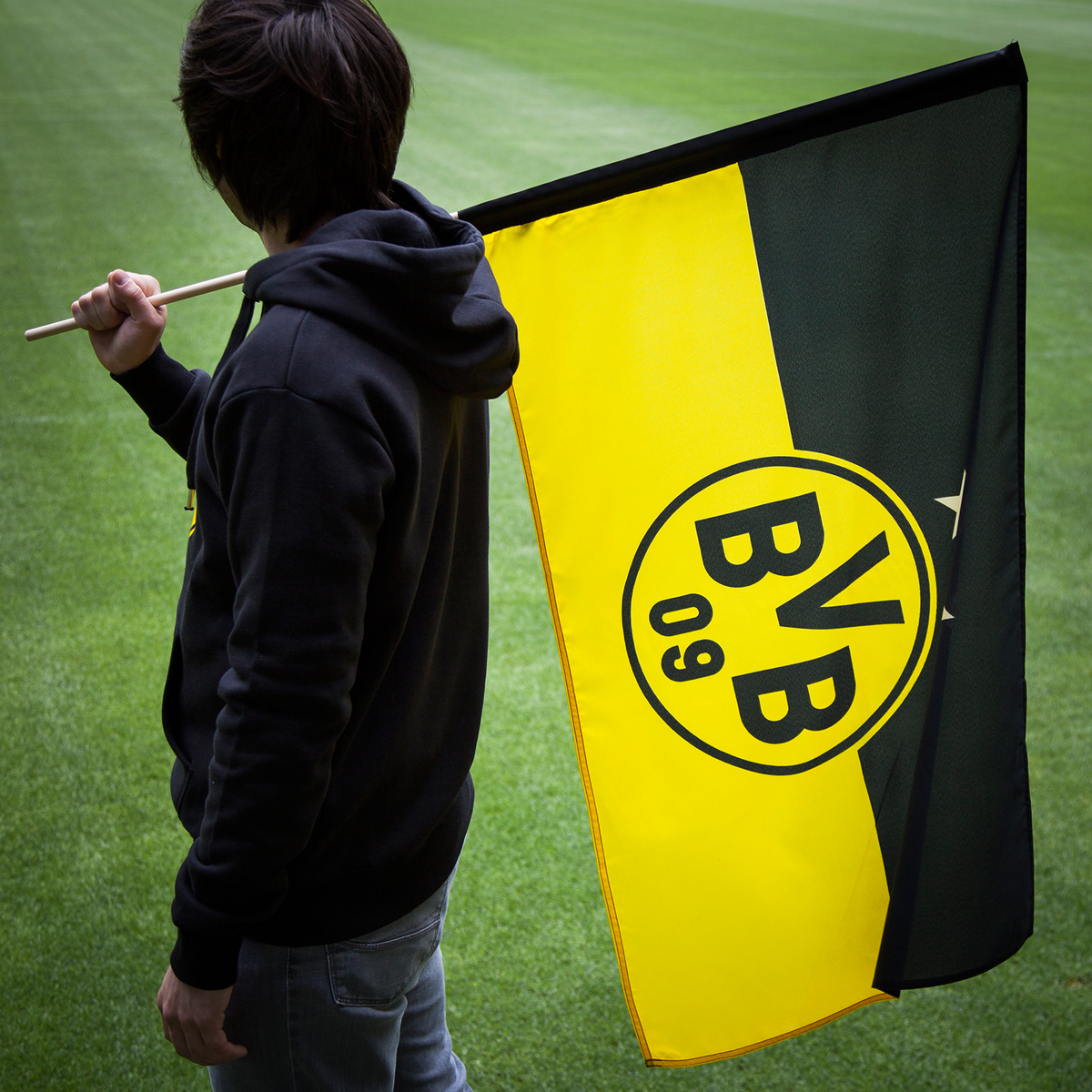Bandera del BVB en negro y amarillo, de 60 x 40 cm, con un gran logotipo central del BVB, sostenida por una persona sobre un campo de fútbol.