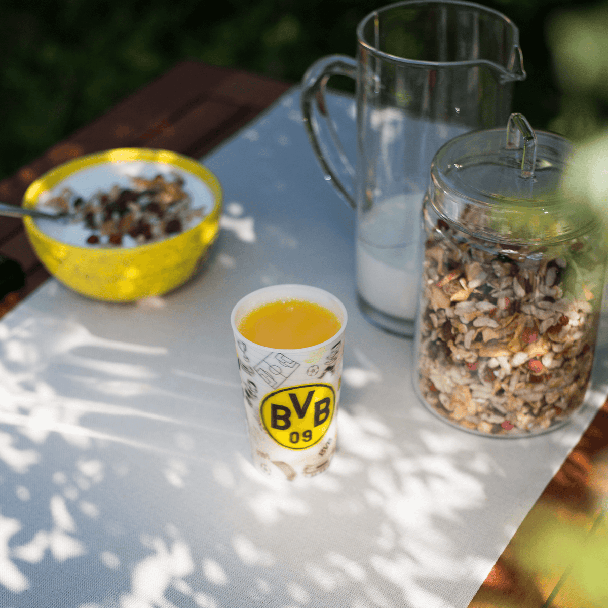 Fan-Glas von Borussia Dortmund mit schwarzgelbem BVB-Logo, gefüllt mit Orangensaft, präsentiert auf einem gedeckten Tisch mit Müslischale, Milchkanne und Vorratsglas.
