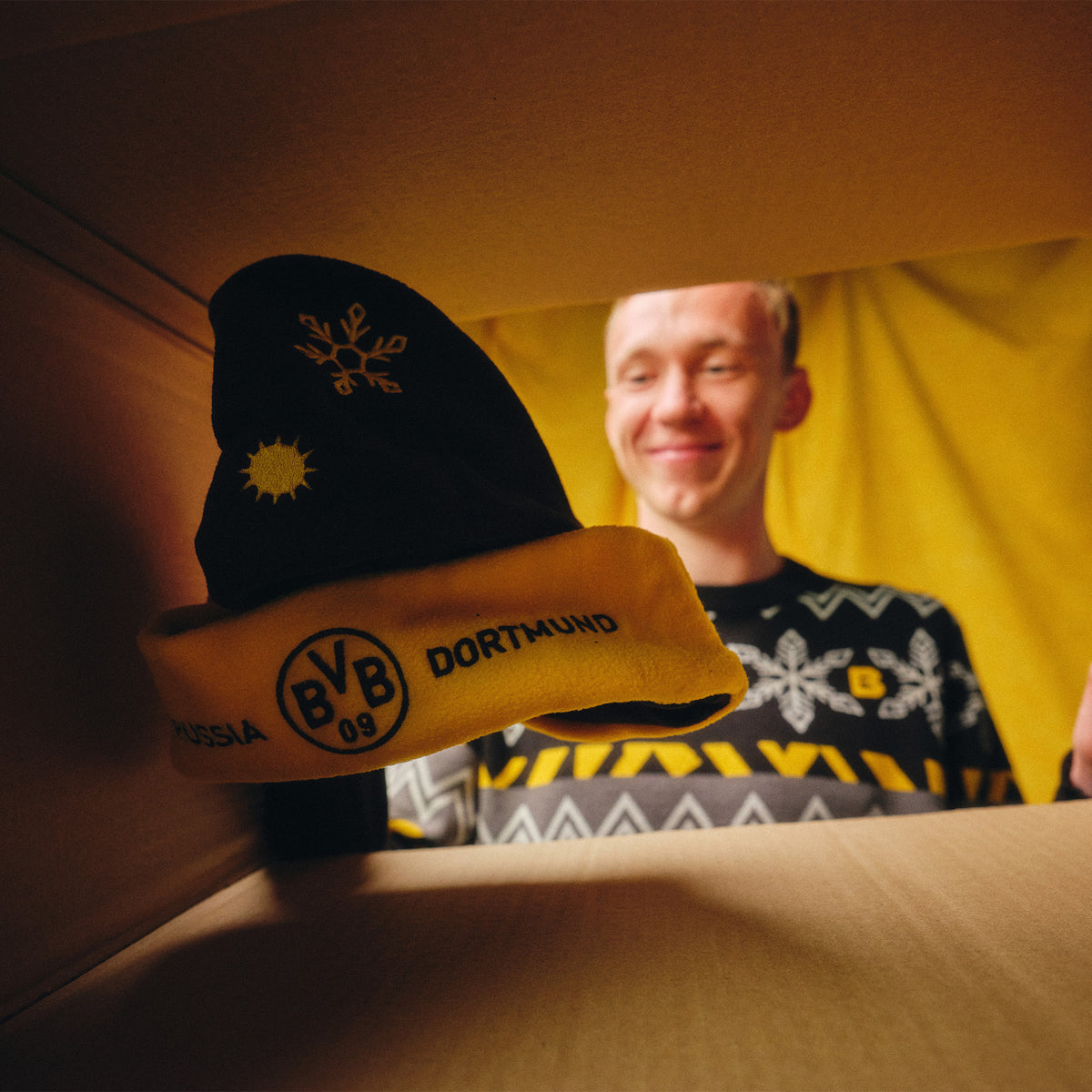 Schwarzgelbe BVB-Weihnachtsmütze mit Schneeflockenmotiv und BVB-Logo, präsentiert von einem Mann in weihnachtlichem Ambiente.
