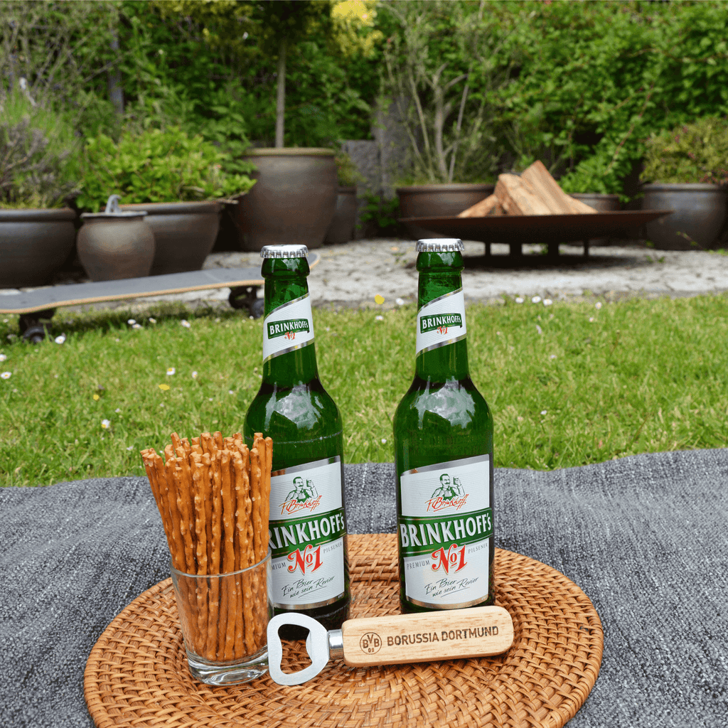 A BVB bottle opener with a wooden handle and the Borussia Dortmund logo, stylishly displayed next to two beer bottles and pretzel sticks during a picnic in the garden.