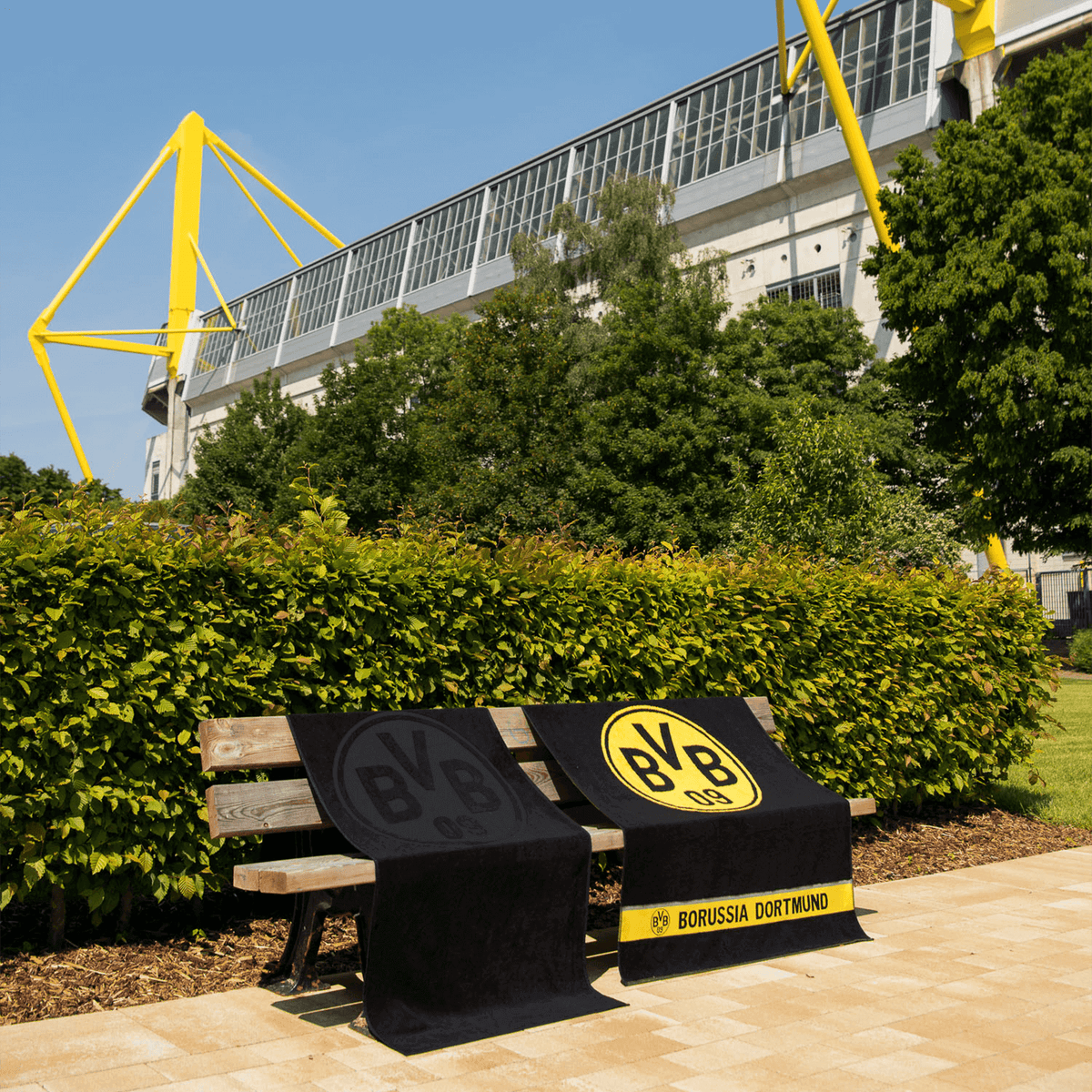 Asciugamani del BVB con design basato sull'emblema: un asciugamano nero con l'emblema nero e un asciugamano con l'emblema giallo e la scritta "Borussia Dortmund", esposti su una panchina davanti al SIGNAL IDUNA PARK.