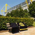 Serviettes du BVB ornées d'un emblème : une serviette noire avec un emblème noir et une serviette avec un emblème jaune et l'inscription « Borussia Dortmund », présentées sur un banc devant le SIGNAL IDUNA PARK.
