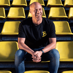 Una maglietta del BVB da uomo, nera e gialla, con il logo della squadra, indossata da un uomo allo stadio seduto su una poltrona gialla.