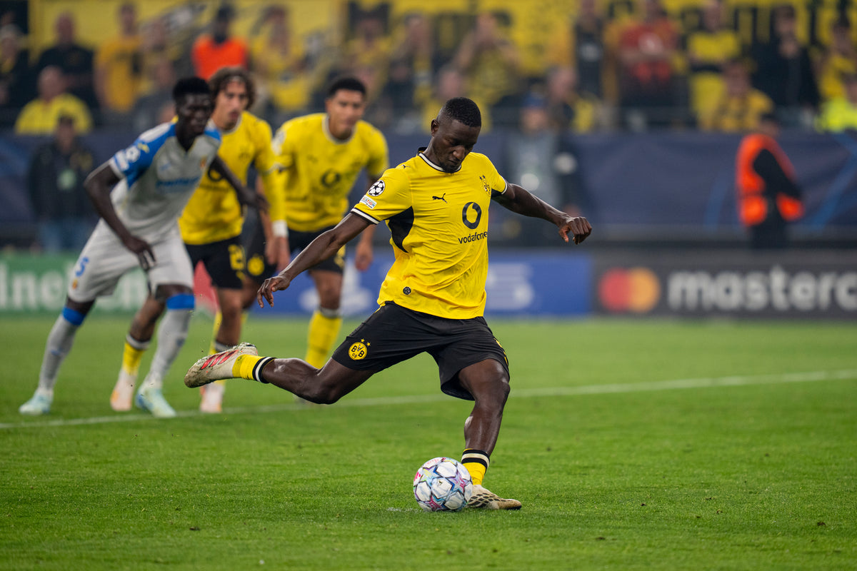 Zu sehen ist der Fußballspieler Serhou Guirassy, der gerade bei einem BVB-Heimspiel im SIGNAL-IDUNA-PARK einen Freistoß schießt. Er trägt das BVB Authentic Heimtrikot der Saison 26/27.