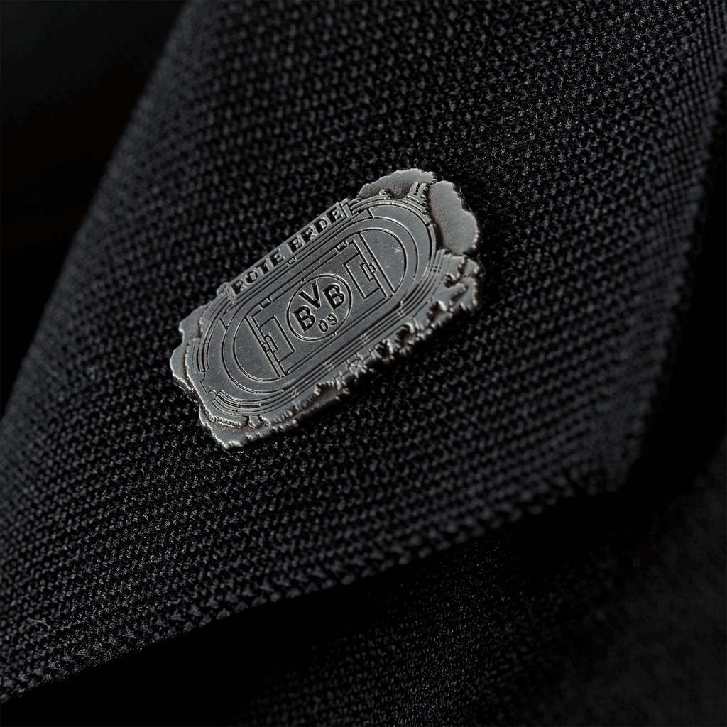 Silberfarbener Borussia-Dortmund-Pin mit Rote-Erde-Stadionumrissen und BVB-Logo, präsentiert in einer Detailaufnahme auf schwarzem Stoff.