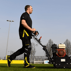 Mitarbeiter mit Borussia Dortmund schwarzem Poloshirt und schwarzgelber Arbeitskleidung beim Bedienen einer Rasenmähmaschine auf einem Fußballplatz des Vereins.