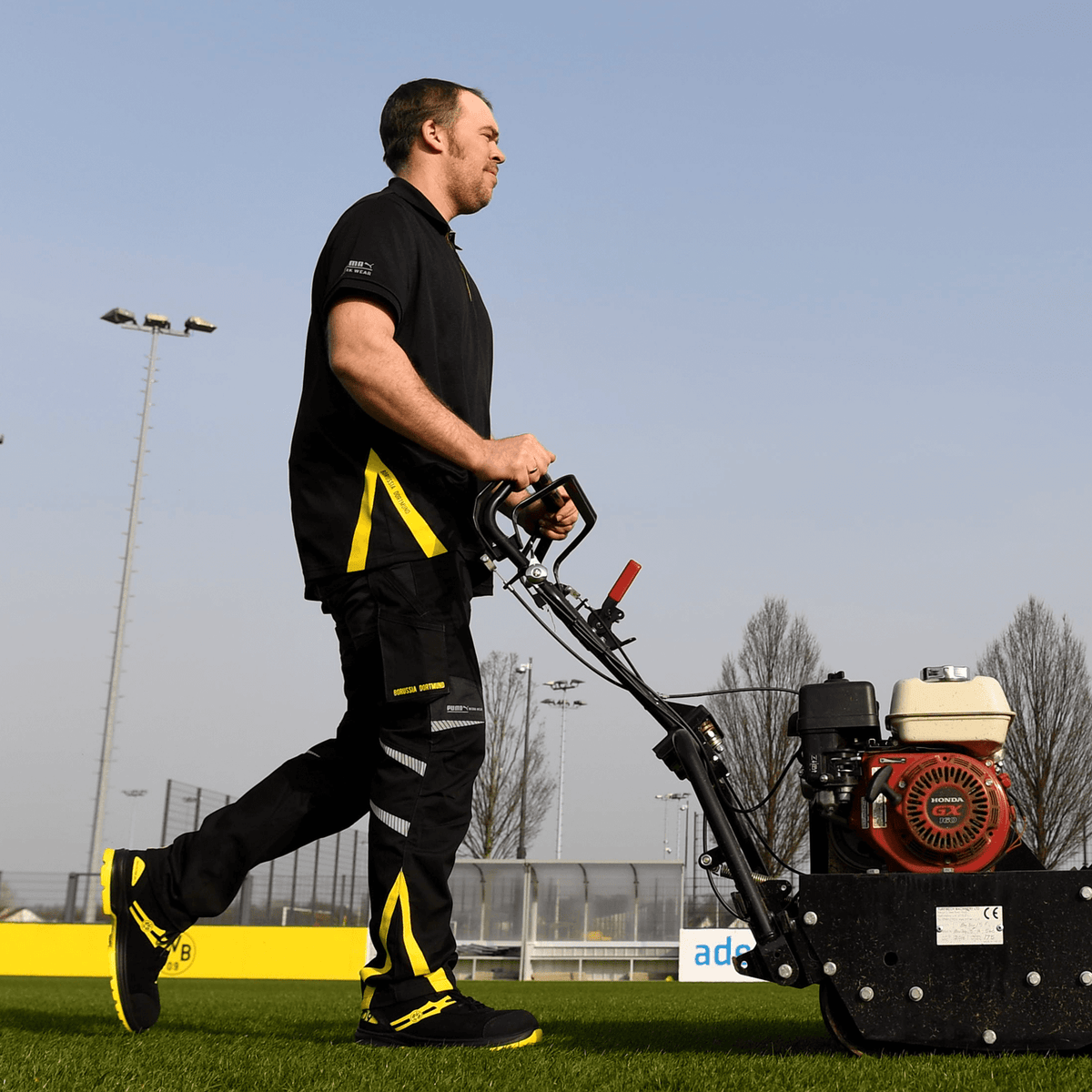 Mitarbeiter mit Borussia Dortmund schwarzem Poloshirt und schwarzgelber Arbeitskleidung beim Bedienen einer Rasenmähmaschine auf einem Fußballplatz des Vereins.
