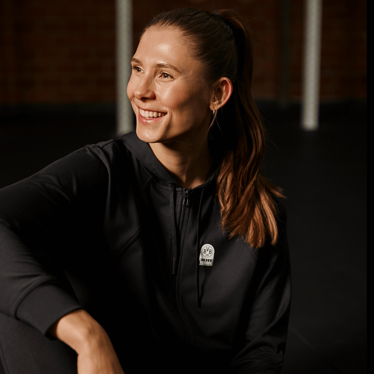 Sweat à capuche zippé noir BVB x PUMA pour femme, orné du logo BVB, présenté par une femme dans un cadre sportif.
