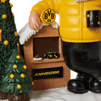 Enano de jardín del BVB con chaqueta amarilla, una taza negra y amarilla, un pequeño puesto de madera con el logotipo del BVB, un árbol de Navidad decorado de forma festiva y un escenario invernal con nieve.