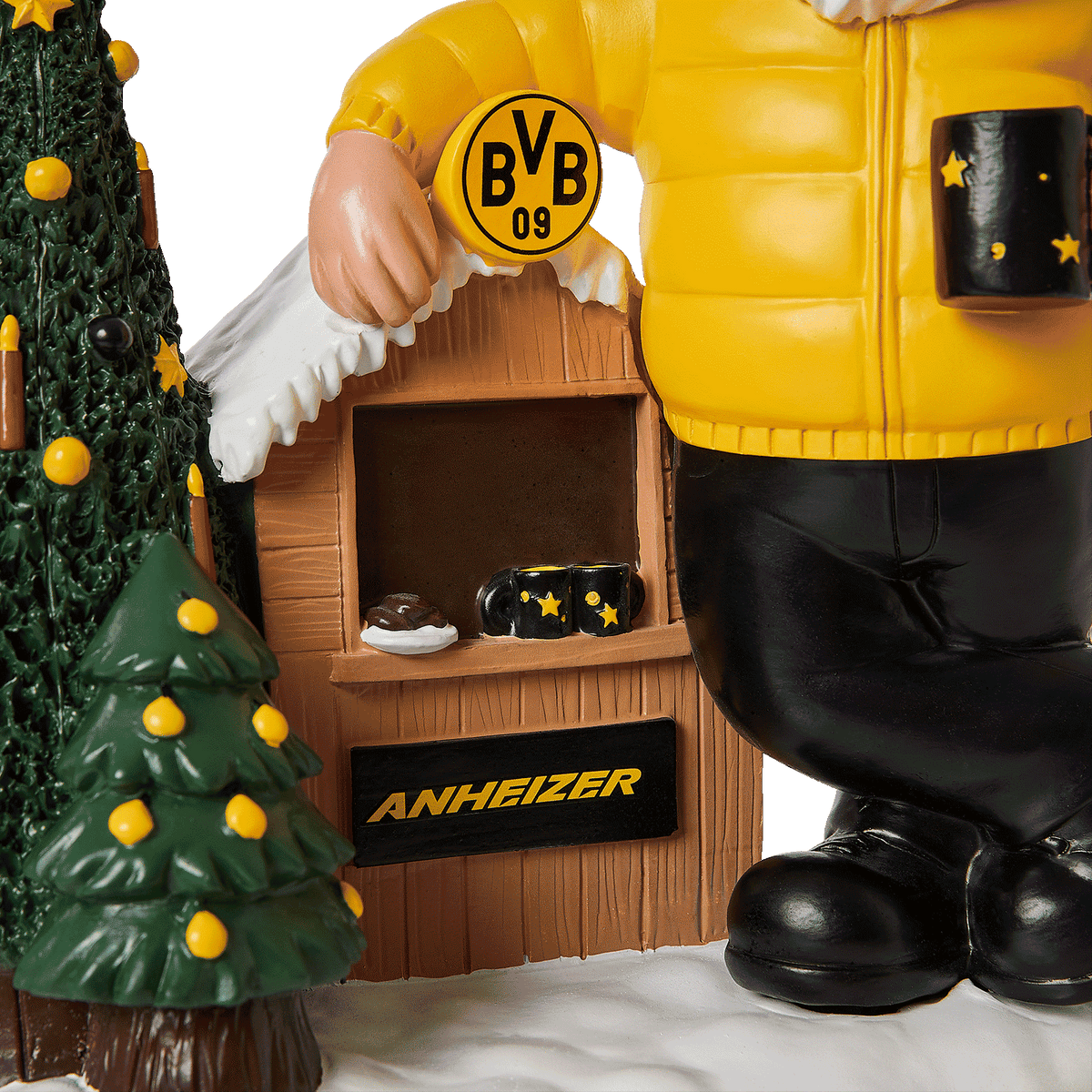 Enano de jardín del BVB con chaqueta amarilla, una taza negra y amarilla, un pequeño puesto de madera con el logotipo del BVB, un árbol de Navidad decorado de forma festiva y un escenario invernal con nieve.