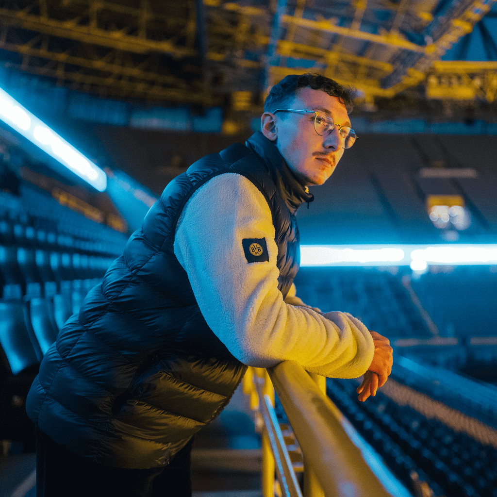 Person trägt eine schwarze BVB Padded Weste mit BVB-Logo und lehnt lässig am Geländer im SIGNAL IDUNA PARK, beleuchtet von Stadionlichtern.