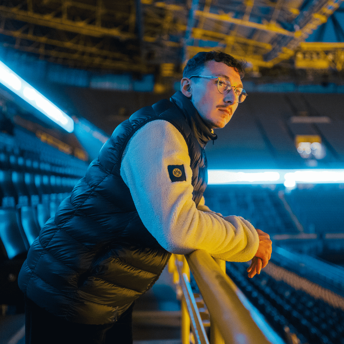 Person trägt eine schwarze BVB Padded Weste mit BVB-Logo und lehnt lässig am Geländer im SIGNAL IDUNA PARK, beleuchtet von Stadionlichtern.