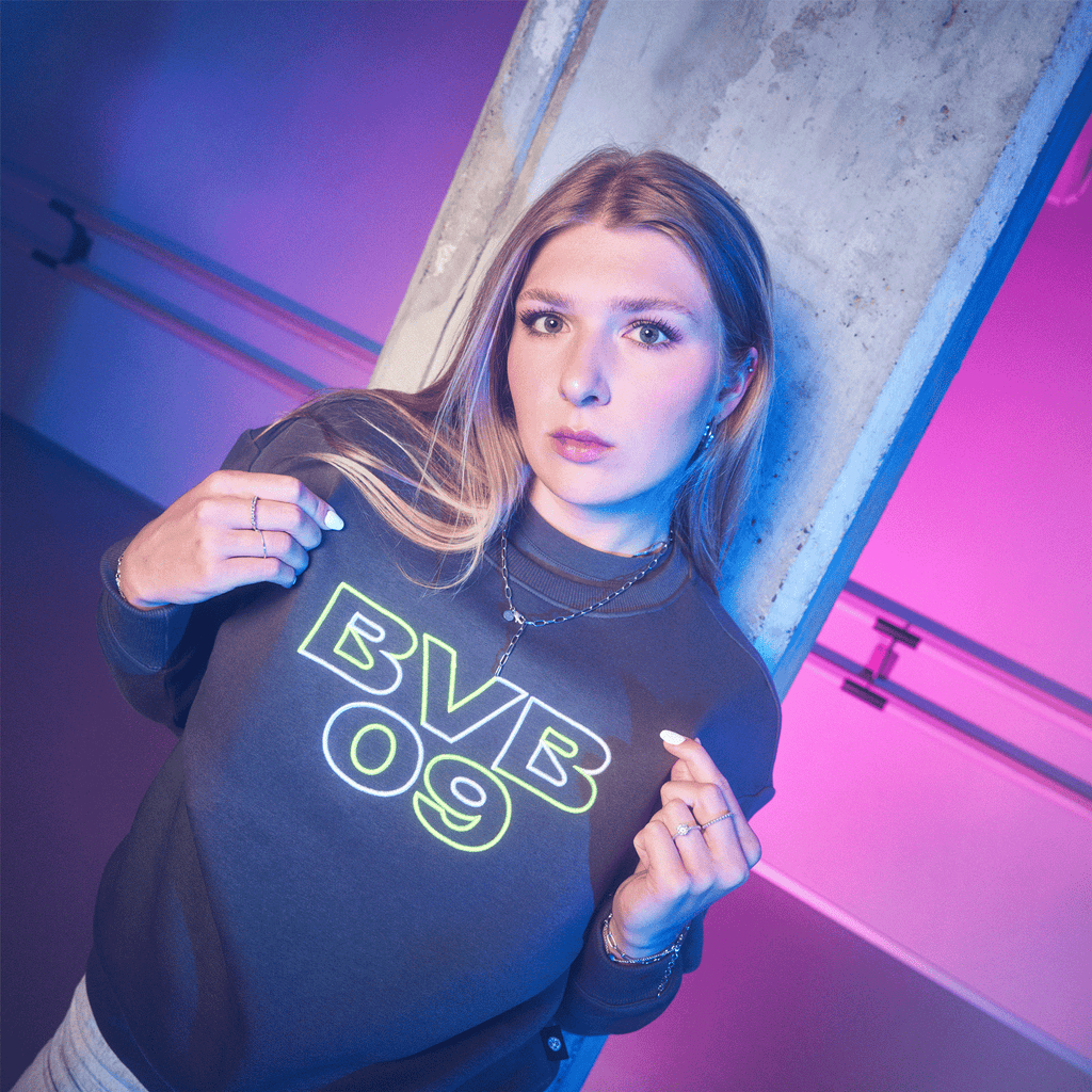 Jersey de cuello redondo negro del BVB para mujer con el logotipo «BVB 09» en colores neón, presentado por una mujer sobre un fondo morado