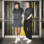 Dos hombres lucen ropa de aficionado del BVB en negro y amarillo, compuesta por una camiseta negra y una sudadera con capucha con detalles amarillos, con un fondo urbano al estilo de una estación de metro.