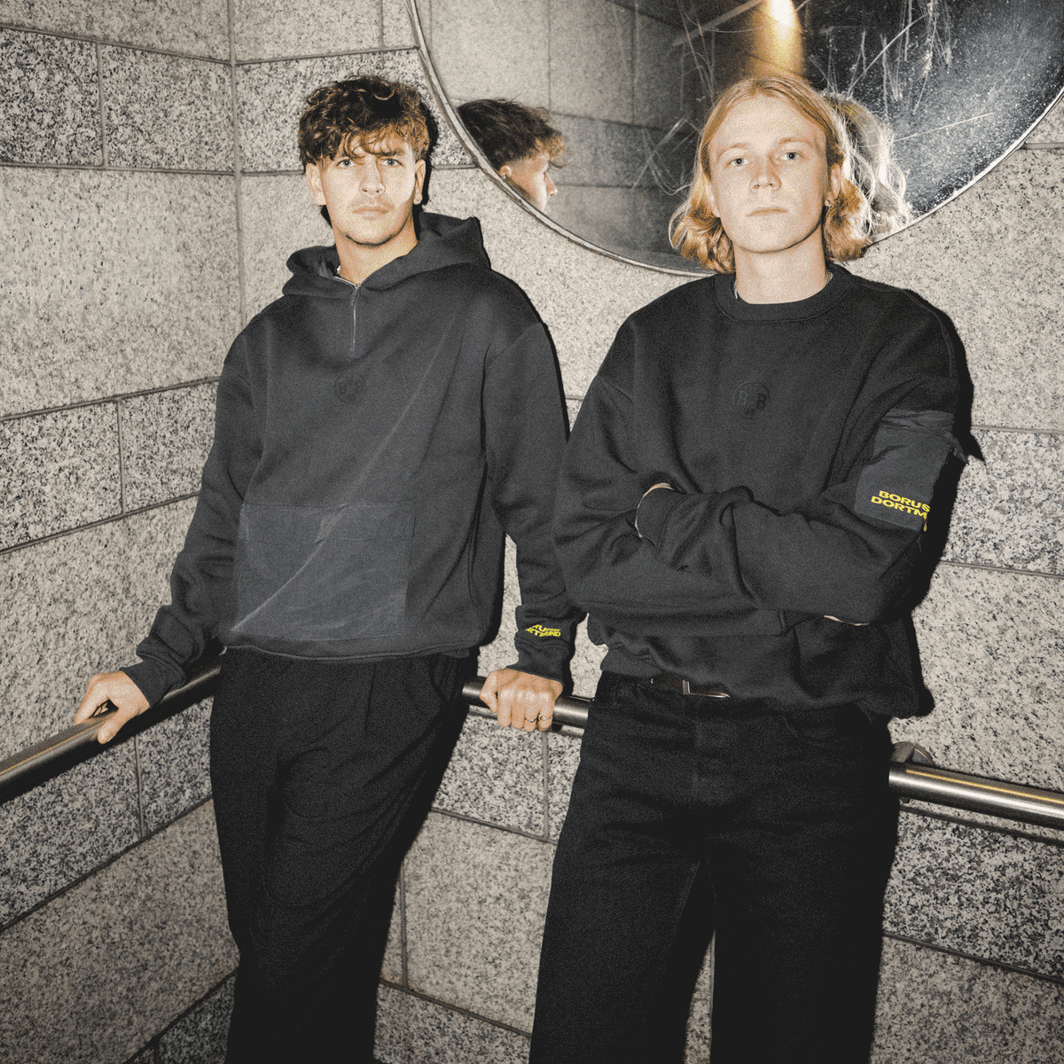 Deux personnes présentent le pull à col rond BVB Underground, de couleur noire et orné de subtils détails aux couleurs du BVB, idéal pour les supporters du Borussia Dortmund qui aiment la mode.