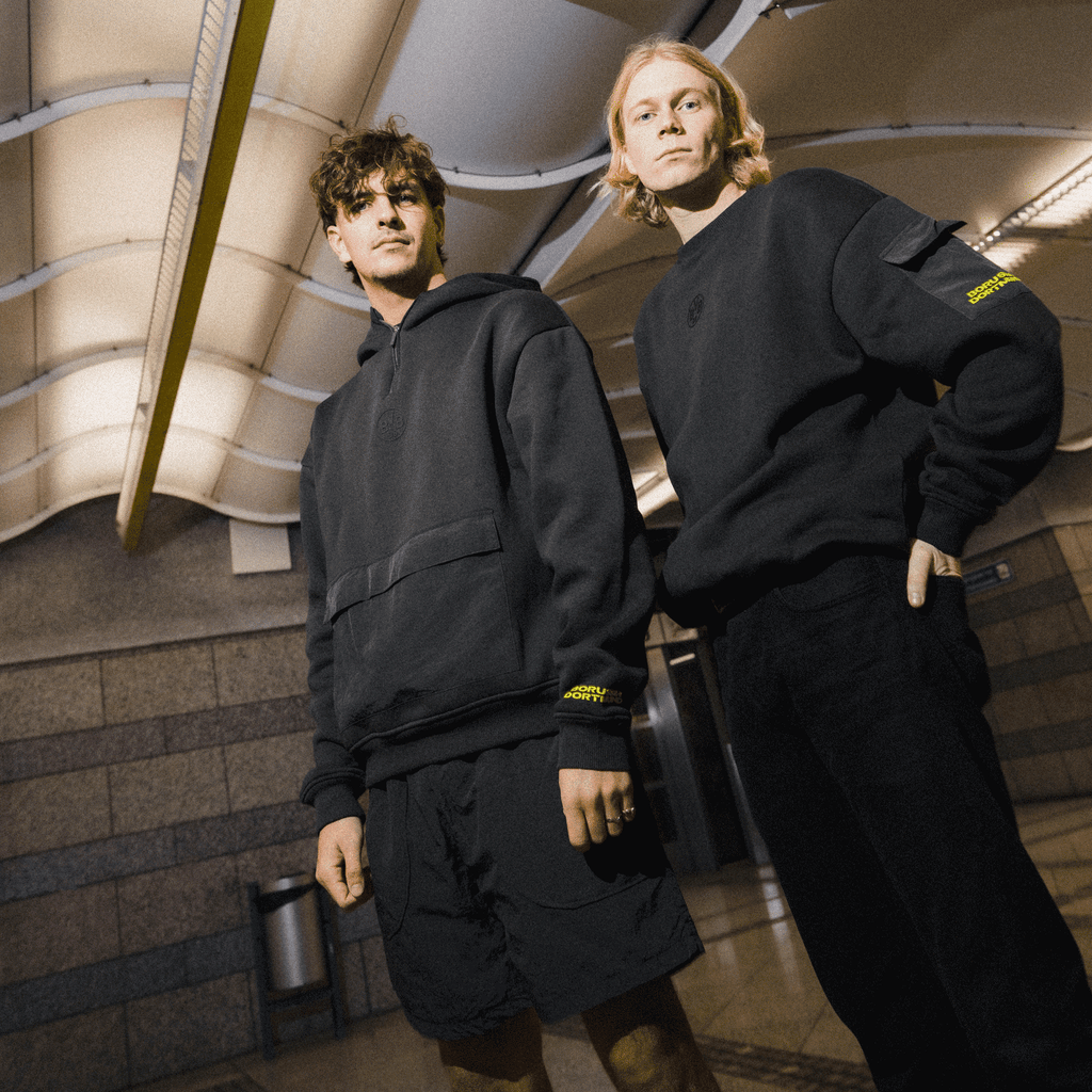 Deux personnes présentent la collection noire de pulls à col rond « BVB Underground » dans un cadre urbain et élégant. Les pulls arborent des détails discrets aux couleurs du BVB et du Borussia Dortmund.