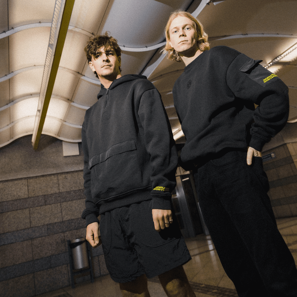 Deux personnes présentent la collection noire de pulls à col rond « BVB Underground » dans un cadre urbain et élégant. Les pulls arborent des détails discrets aux couleurs du BVB et du Borussia Dortmund.