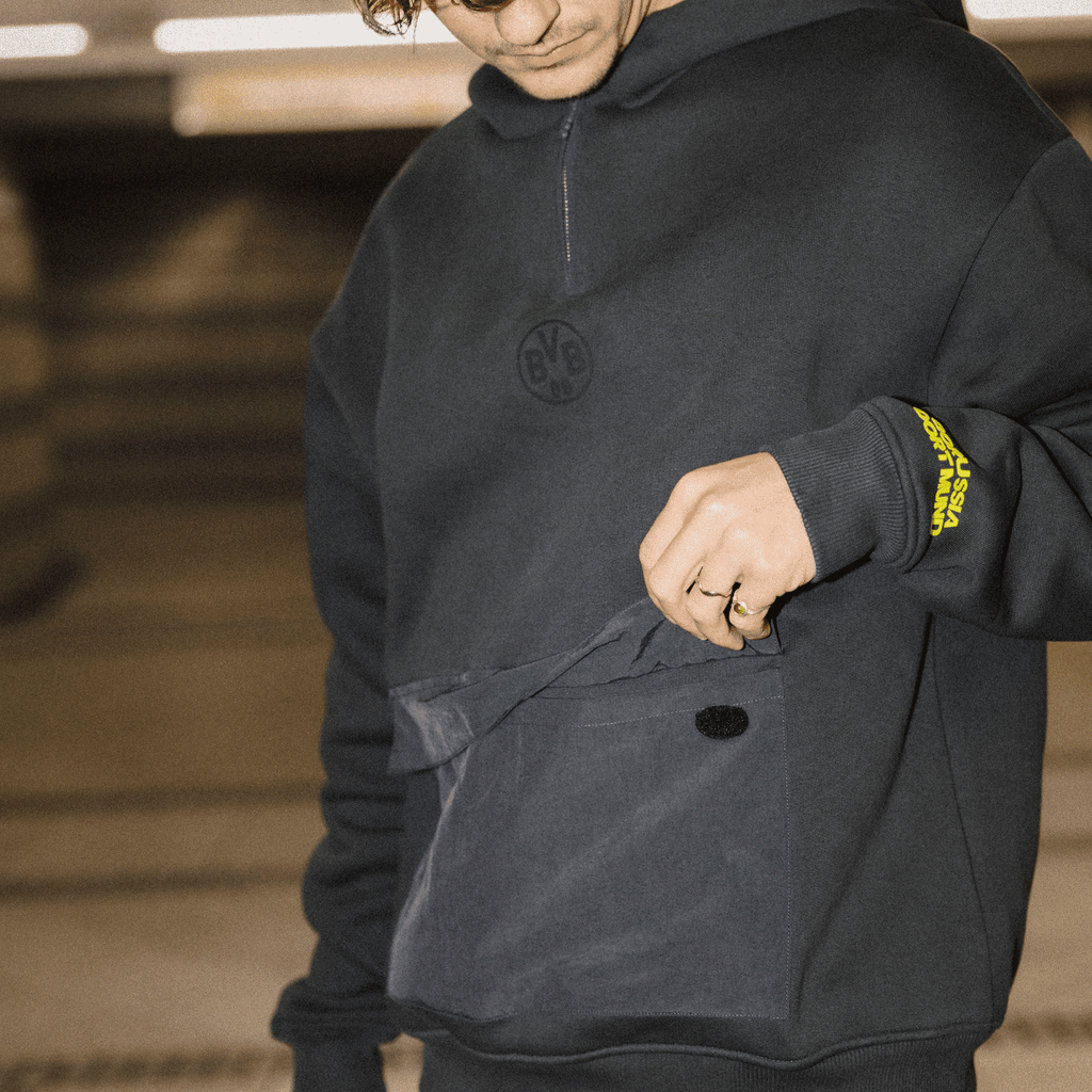 Schwarzer BVB-Hoodie mit Kapuze und logo-geprägten Details, gelber Borussia Dortmund Schriftzug am Ärmel, sportlich und stylisch.