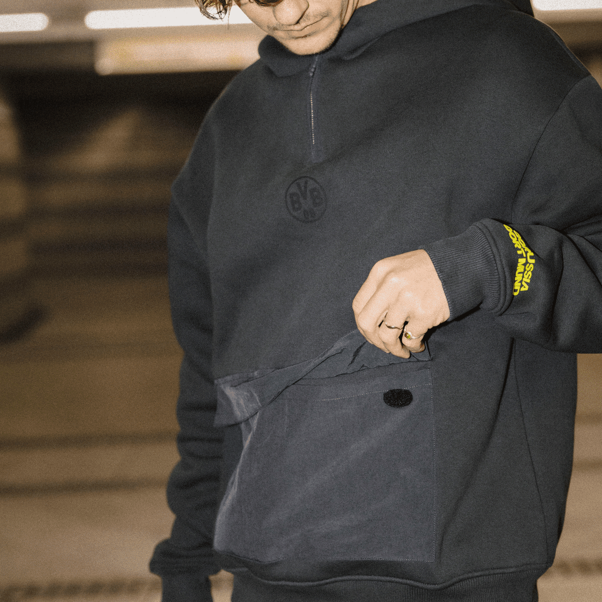 Schwarzer BVB-Hoodie mit Kapuze und logo-geprägten Details, gelber Borussia Dortmund Schriftzug am Ärmel, sportlich und stylisch.