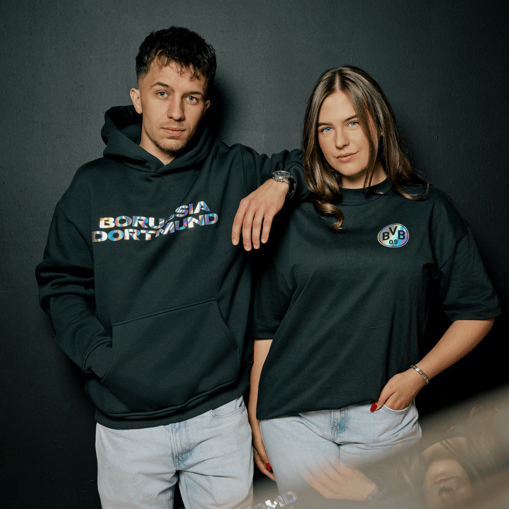 Schwarzer BVB Rainbow Hoodie mit Borussia Dortmund Schriftzug im Regenbogen-Design sowie schwarzes T-Shirt mit BVB 09 Emblem im Regenbogen-Look, präsentiert von zwei Personen.