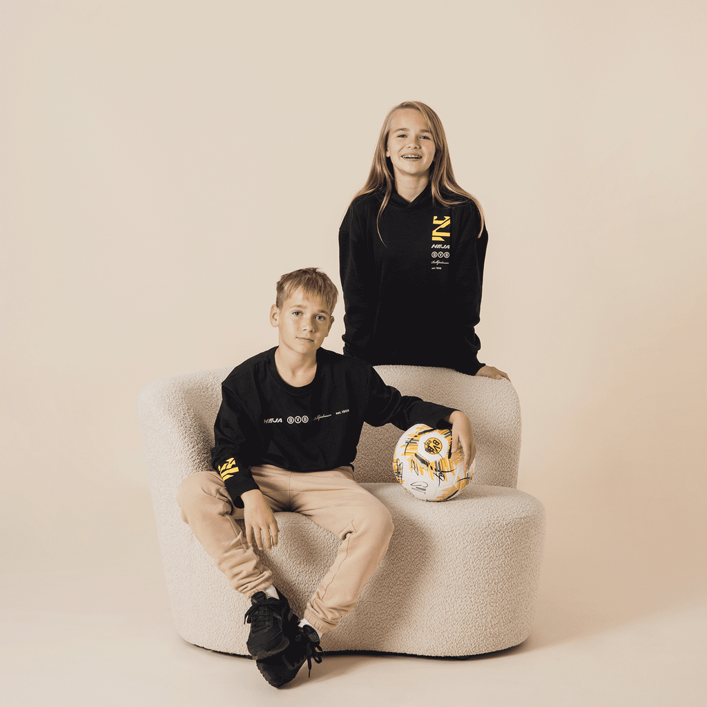 A boy and a girl are modeling BVB fan apparel for kids, consisting of a black oversized long-sleeve shirt and a hoodie with yellow details.