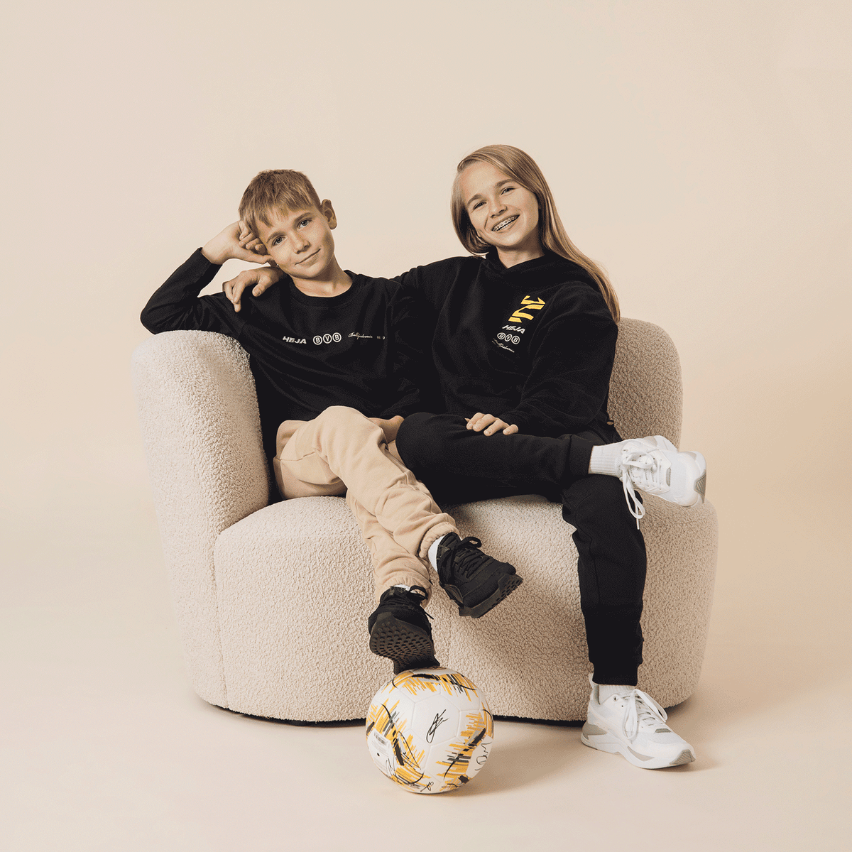 Zwei Kinder in schwarzen BVB Longsleeves, lässig auf einem Sessel sitzend, mit einem BVB-Fußball im Vordergrund, präsentiert als Fanartikel.