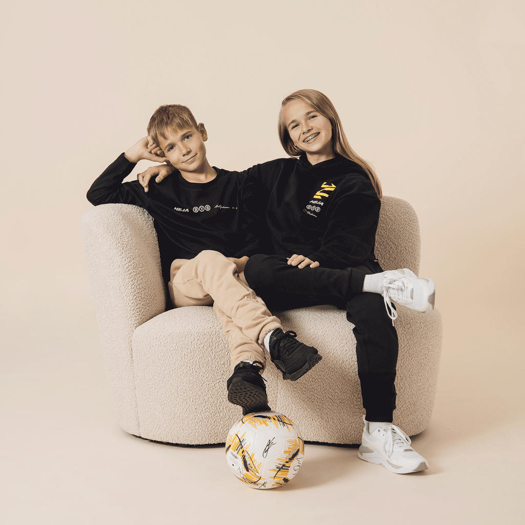 Two children wearing black BVB long-sleeve shirts, sitting casually in an armchair, with a BVB soccer ball in the foreground, presented as fan merchandise.