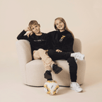 Two children wearing black BVB long-sleeve shirts, sitting casually in an armchair, with a BVB soccer ball in the foreground, presented as fan merchandise.