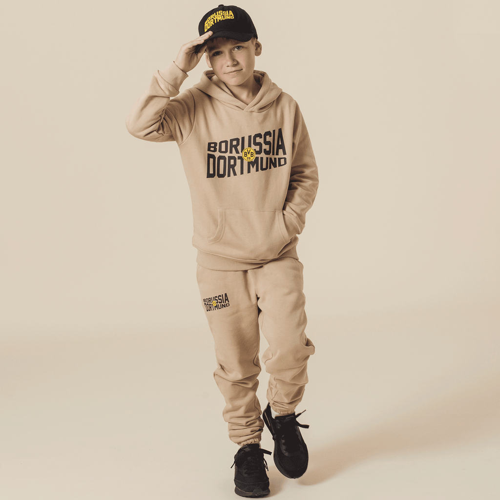 L'immagine mostra un bambino che indossa pantaloni della tuta beige del BVB e una felpa con cappuccio con il logo del Borussia Dortmund, oltre a un cappellino nero e giallo del BVB, adatto al tempo libero e ai tifosi.