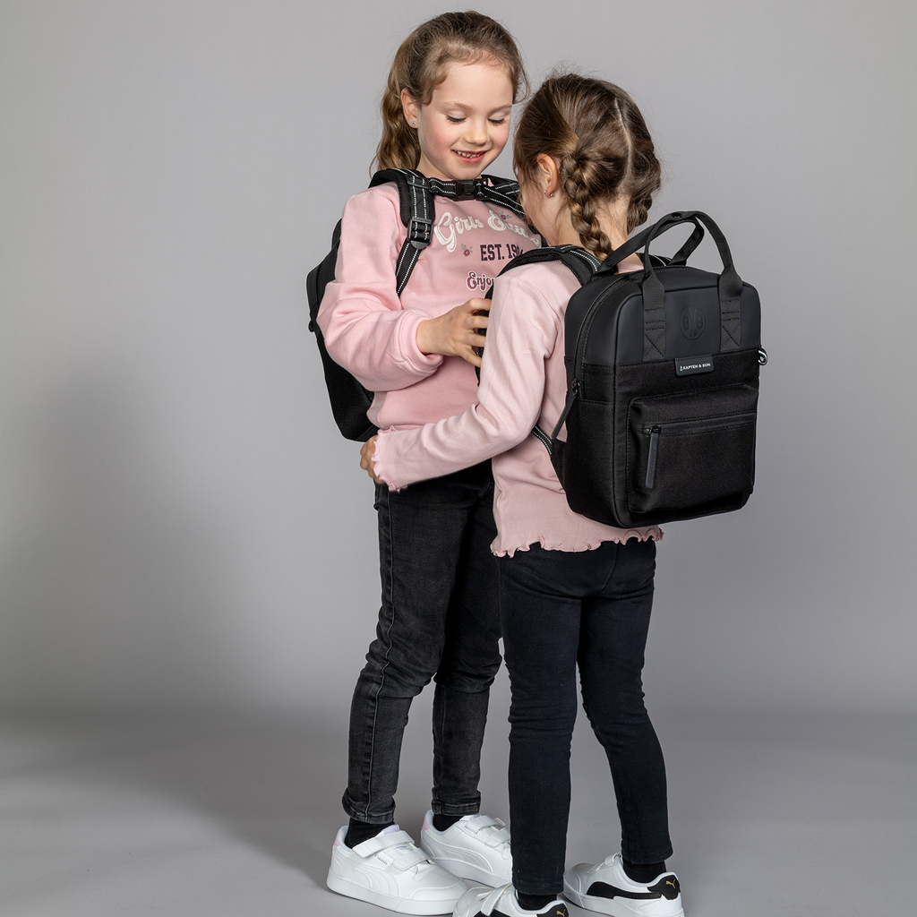 Zwei Kinder tragen schwarze BVB Kapten & Son Rucksäcke, stehen sich lächelnd gegenüber und präsentieren das Produkt in einer Studioaufnahme.