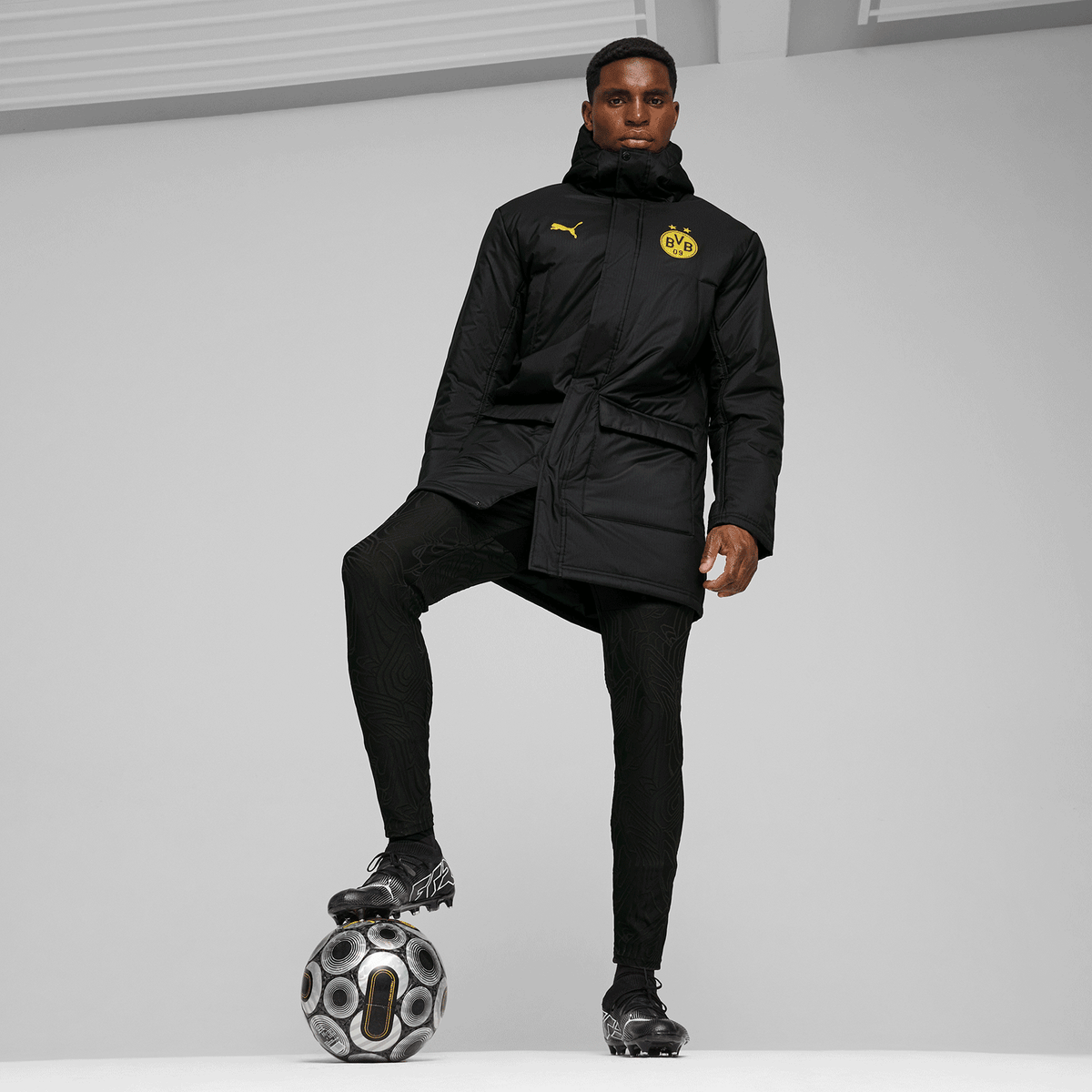 Giacca invernale nera del BVB della collezione 24/25 con stemma del BVB e logo PUMA, indossata da un uomo con un pallone da calcio.