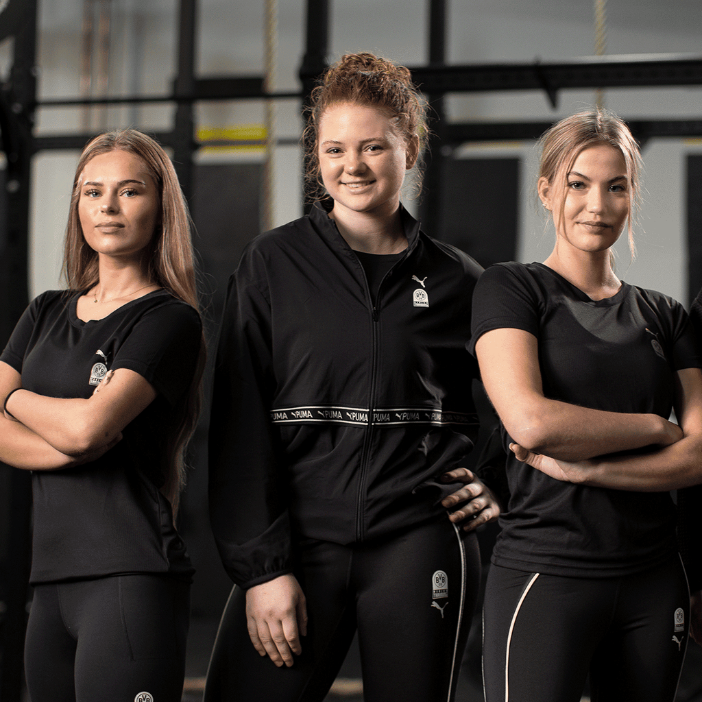 Die BVB x Puma Gym Woven Elastic Damenjacke in elegantem Schwarz, getragen von drei Frauen in einem Fitnessstudio. Ideal für Sport und Freizeit.