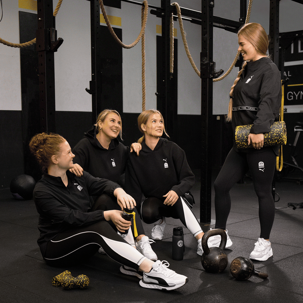 Sportliche Damenmode der BVB x Puma Kollektion, präsentiert von vier Frauen in einem Fitnessstudio mit Kettlebells und BVB-Motiv.