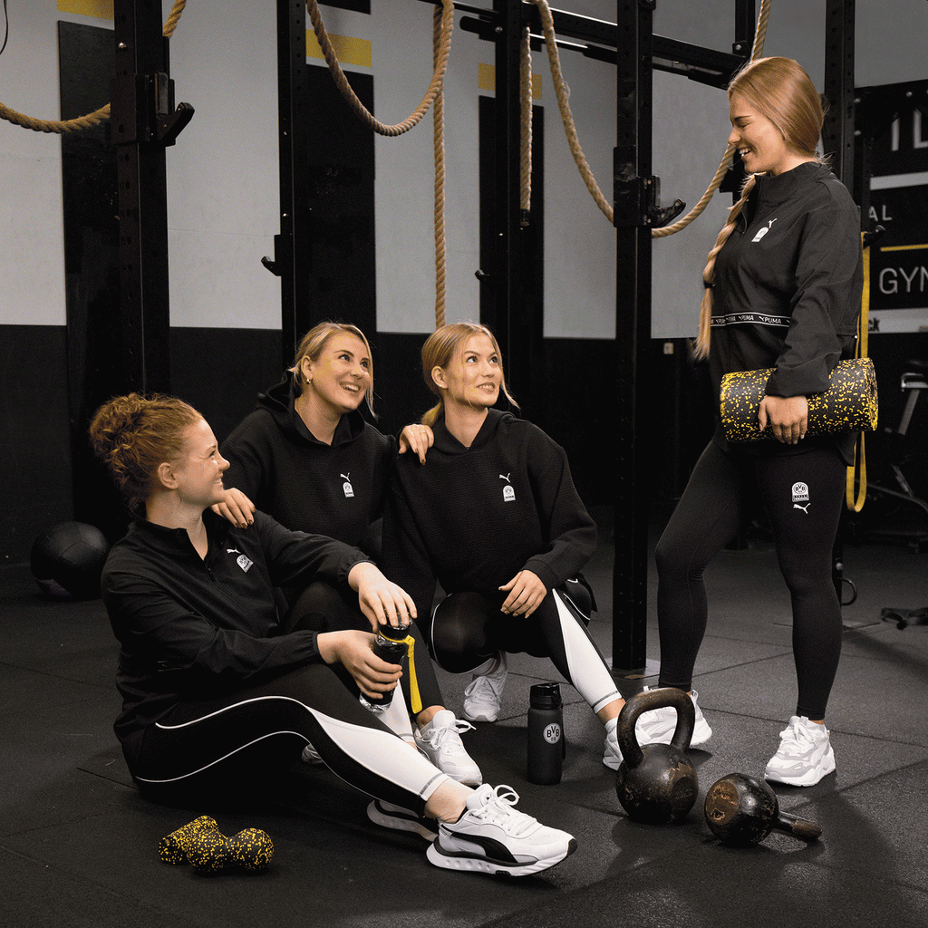 スポーツホールで、4人の女性が、BVB x Pumaの「Gym Train Strong 7/8 Tight（レディース）」と、BVBおよびPumaのロゴが入った黒いフーディを着用し、ケトルベルやスポーツ器具に囲まれている。