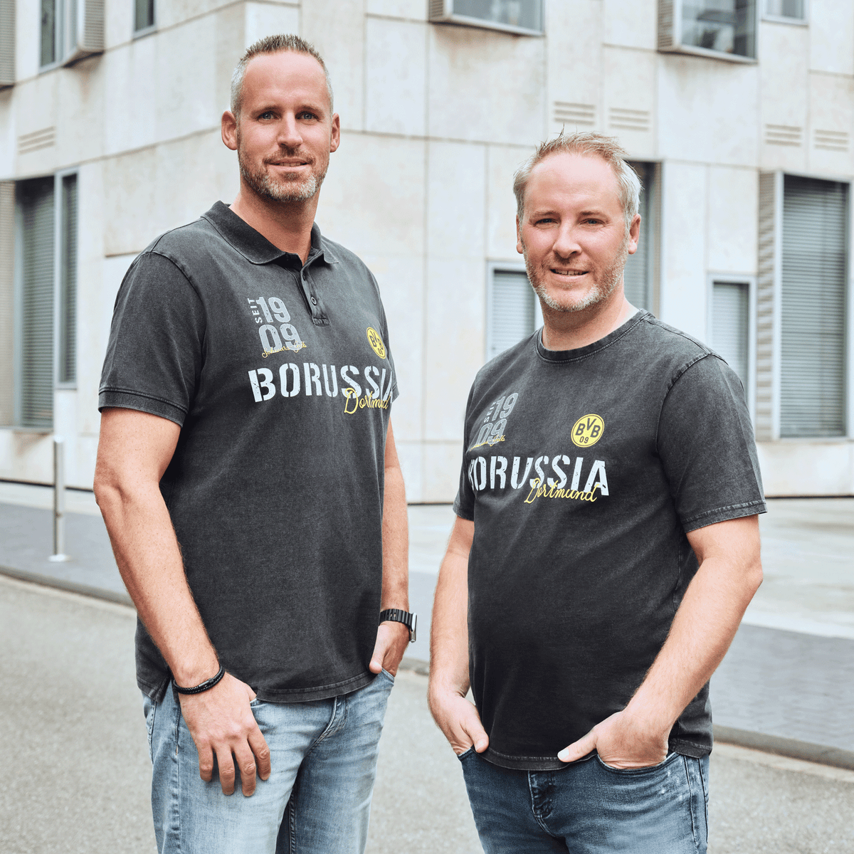 Zwei Männer in schwarzgrauen BVB-Shirts mit Borussia-Dortmund-Schriftzug und 1909-Aufdruck, stehend vor einem modernen Gebäude.