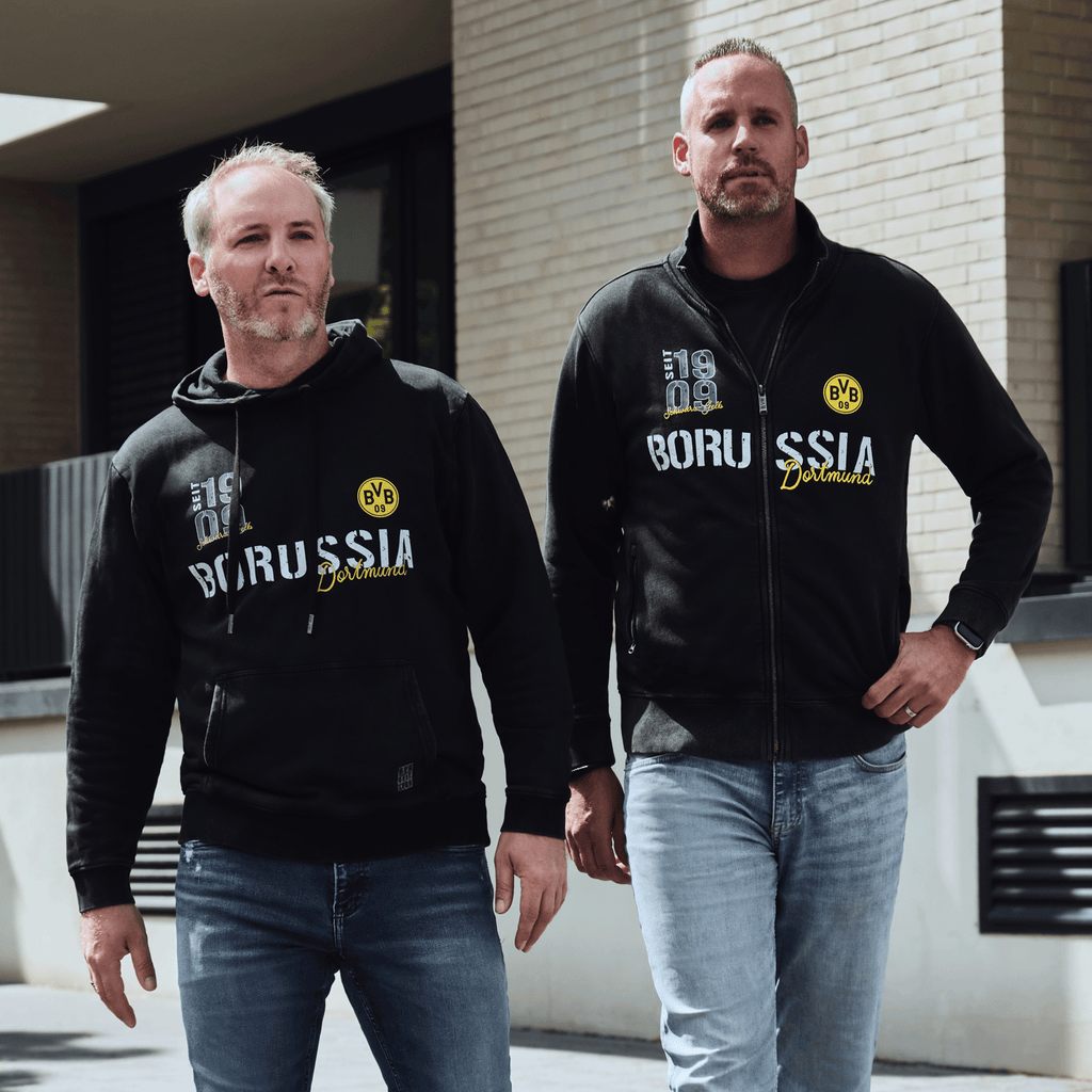 Zwei Männer präsentieren einen schwarzen BVB-Hoodie und eine schwarze BVB-Jacke mit gelbem Borussia Dortmund-Logo und Schriftzug, ideal für Freizeit und Fanmode.