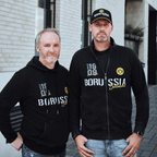 两名男子展示着多特蒙德（BVB）的球迷周边商品：一件黑色拉链夹克和一件连帽衫，上面印有黄色和白色的“1909”和“多特蒙德”字样，以及多特蒙德的队徽。