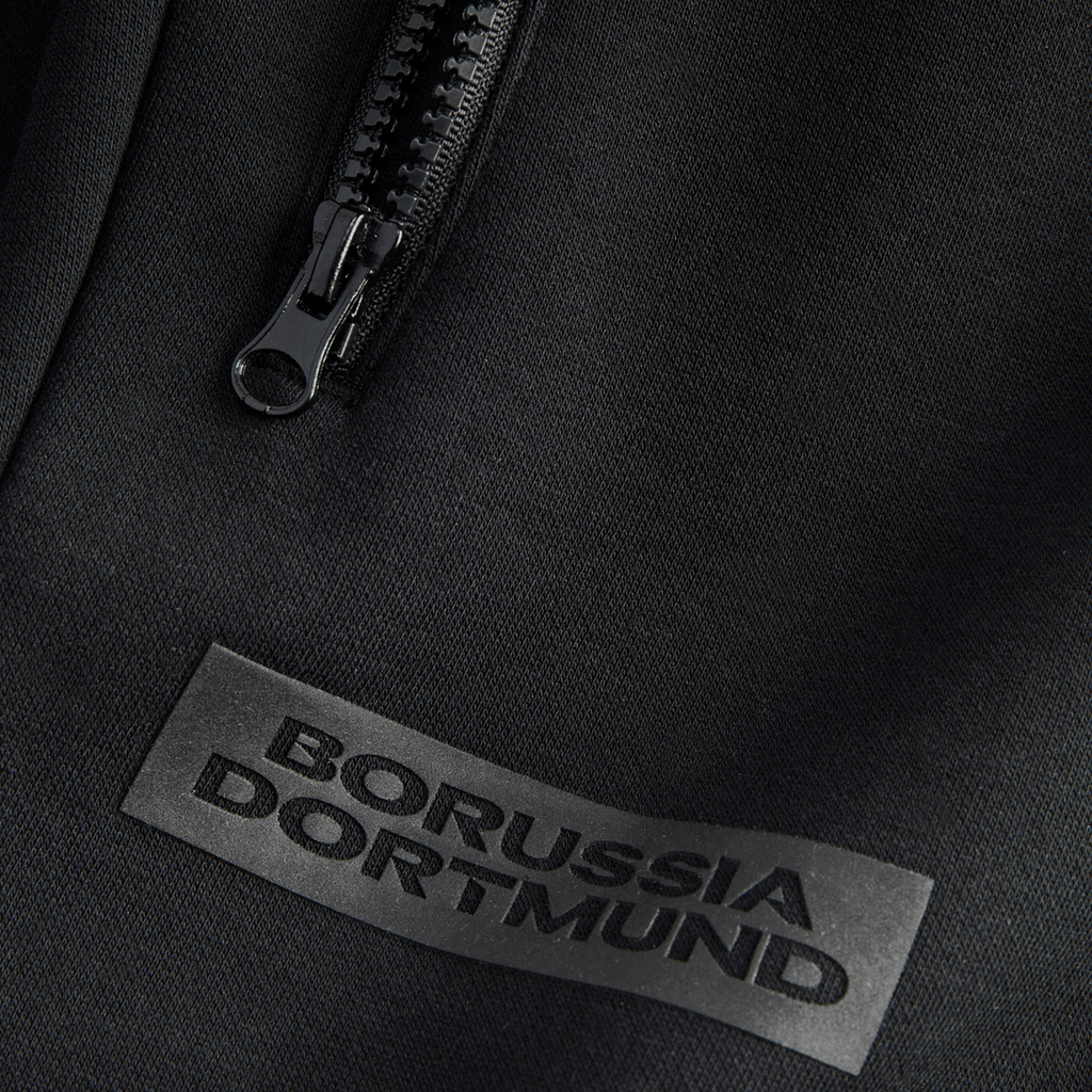 Pantalones de chándal negros «BVB Essentials» con el logotipo del Borussia Dortmund y cremallera, mostrados en detalle.