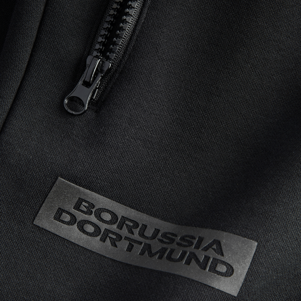 Pantalones de chándal negros «BVB Essentials» con el logotipo del Borussia Dortmund y cremallera, mostrados en detalle.