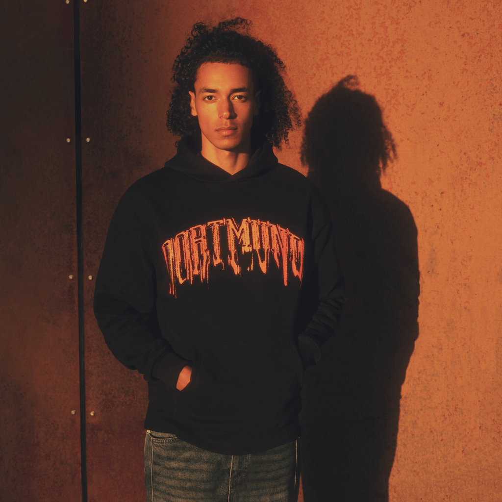 Sudadera negra con capucha del BVB con el llamativo logotipo de Dortmund en naranja en la parte delantera, lucida por una persona en una pose desenfadada ante un fondo con una iluminación cálida.