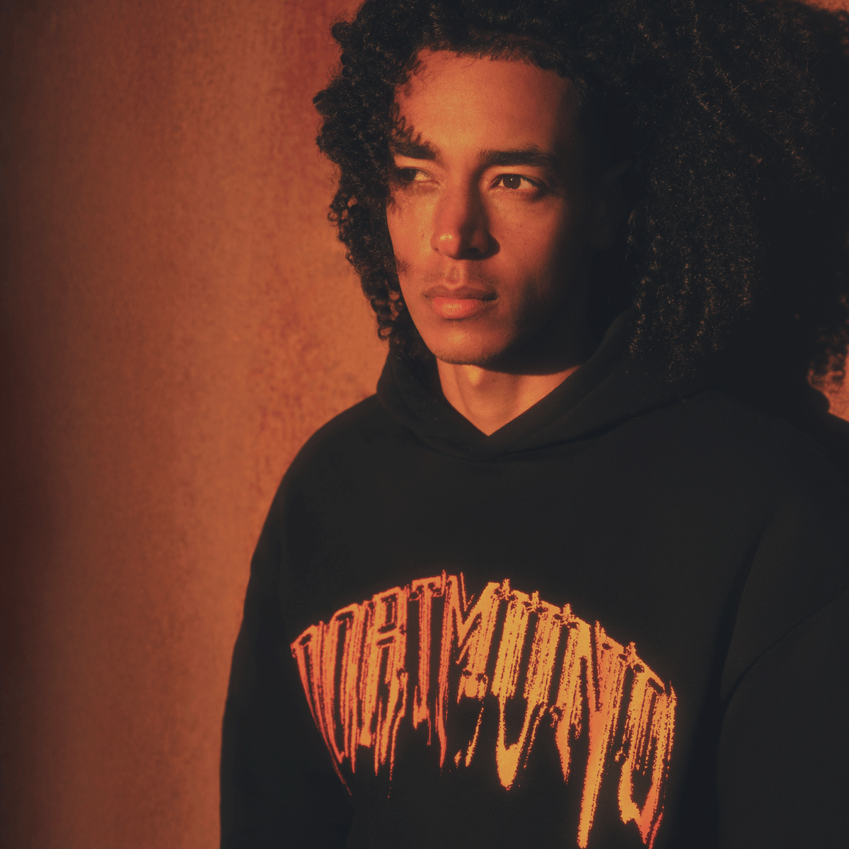 Sweat à capuche noir du BVB orné d'une inscription « Dortmund » orange bien visible, présenté par une personne sous un éclairage évocateur.
