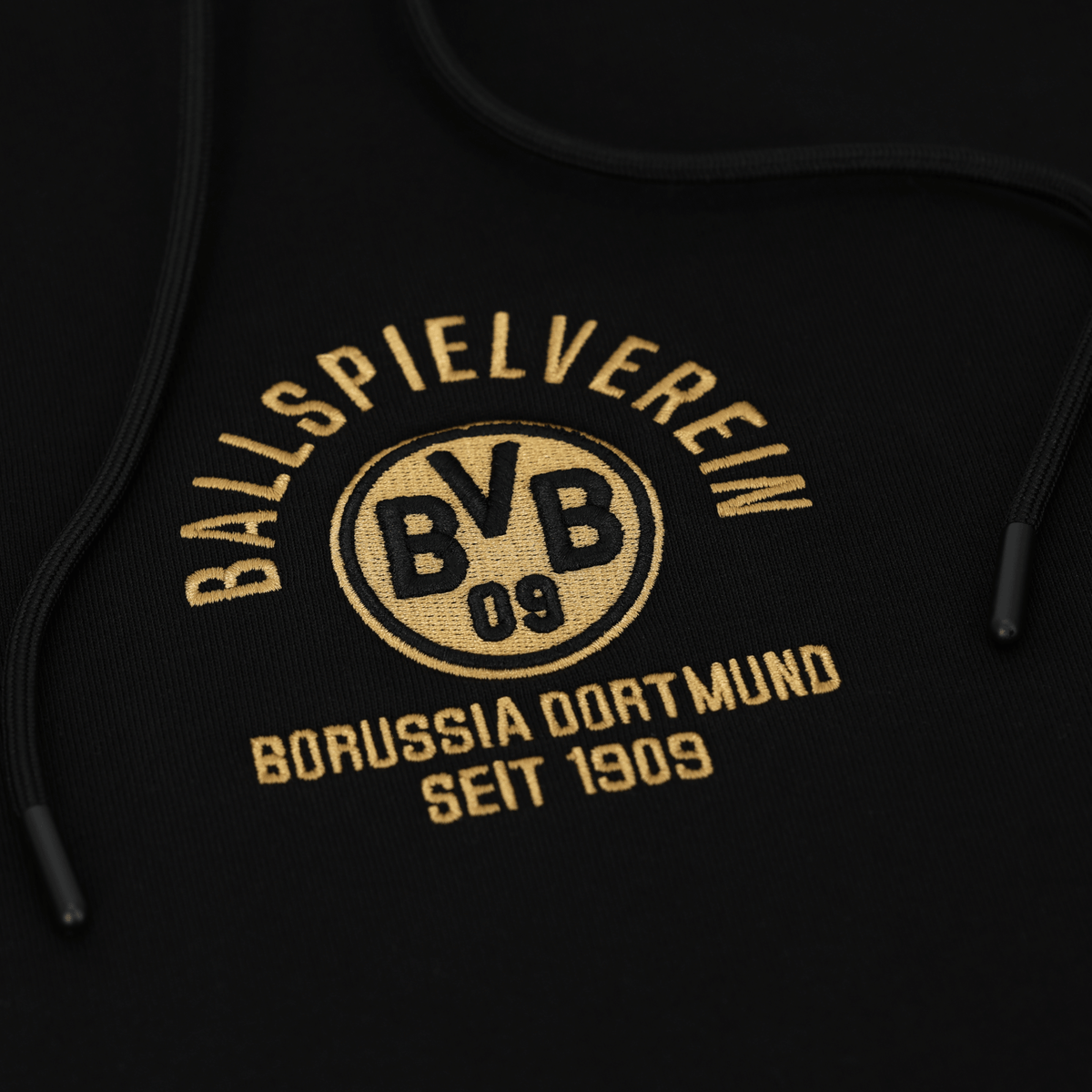 Primo piano di una felpa con cappuccio nera del BVB con la scritta dorata "Ballspielverein Borussia Dortmund seit 1909" e il logo del BVB.
