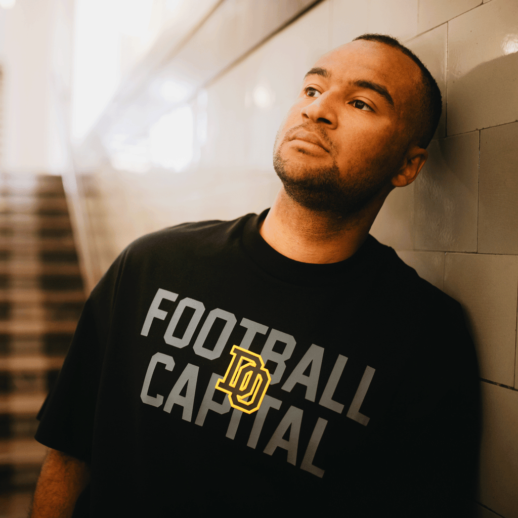 T-shirt BVB College noir avec l'inscription « FOOTBALL CAPITAL » en gris et jaune, porté par une personne ; idéal comme article de fan pour les supporters du Borussia Dortmund, dans un style sportif et décontracté
