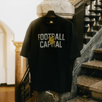 Camiseta negra del BVB College 2025 con el logotipo amarillo y la inscripción gris «Football Capital», expuesta en una percha junto a una escalera.