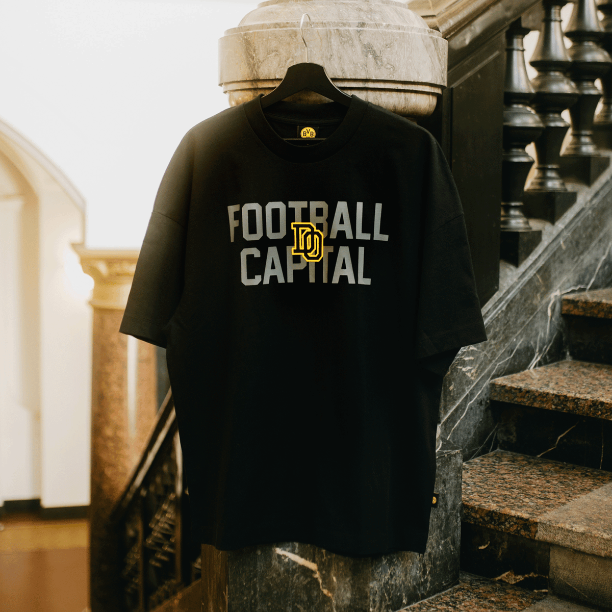 Camiseta negra del BVB College 2025 con el logotipo amarillo y la inscripción gris «Football Capital», expuesta en una percha junto a una escalera.