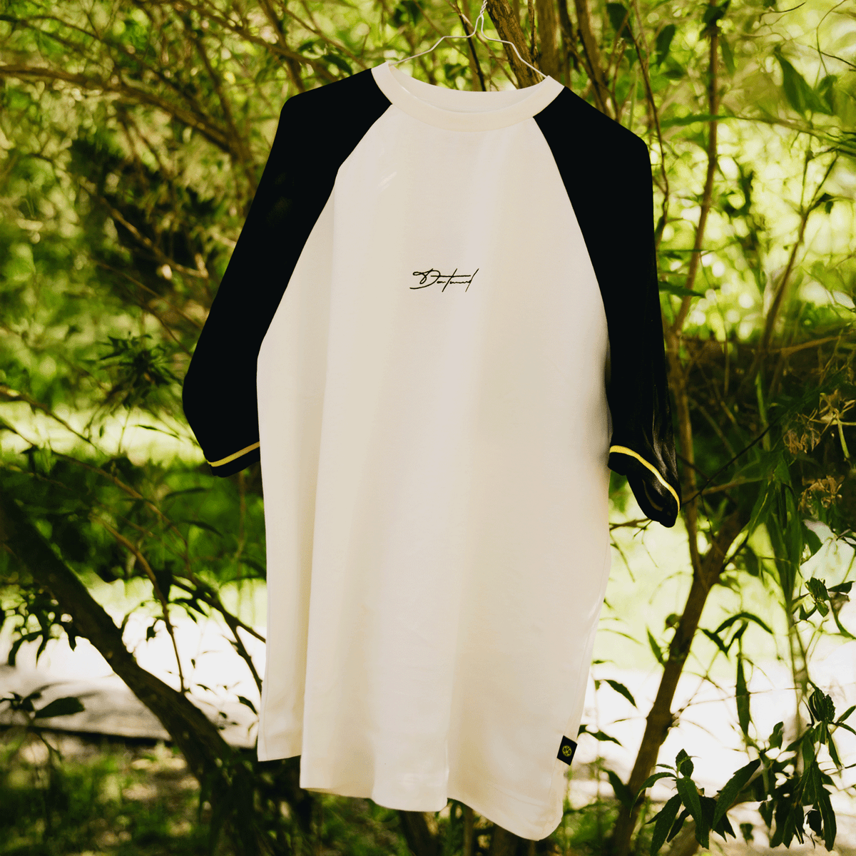 BVB Iconic Lifestyle T-shirt in off-white with black three-quarter sleeves, a yellow trim along the cuffs, and a simple logo print, presented against a natural landscape backdrop.