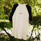 BVB Iconic Lifestyle T-Shirt in Offwhite mit schwarzen Dreiviertelärmeln, gelber Zierlinie an den Bündchen und schlichtem Logo-Print, präsentiert vor einer natürlichen Hintergrundlandschaft.