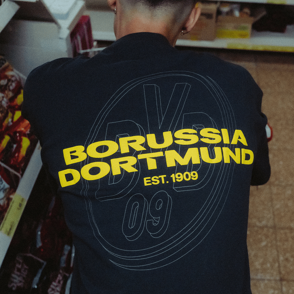 黄色のボルシア・ドルトムントのロゴと「EST. 1909」の文字が入った、黒色のBVBアーバンTシャツの背面。2025年シーズンのファンアイテム。