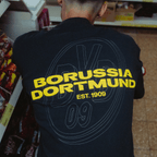 Vista posteriore di una maglietta nera BVB Urban con il logo giallo del Borussia Dortmund e la scritta “EST. 1909”, articolo per i tifosi della stagione 2025.