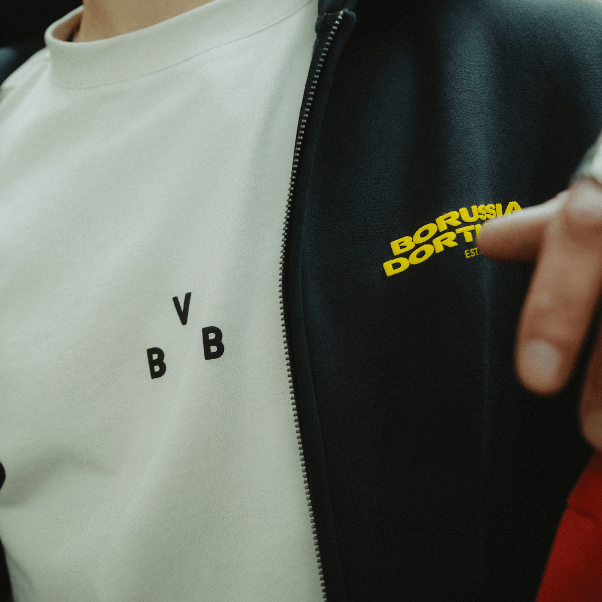 Beiges BVB Urban Tee 2025 mit schwarzem BVB-Logo, kombiniert mit einer schwarzen Jacke mit gelbem Borussia Dortmund Schriftzug.